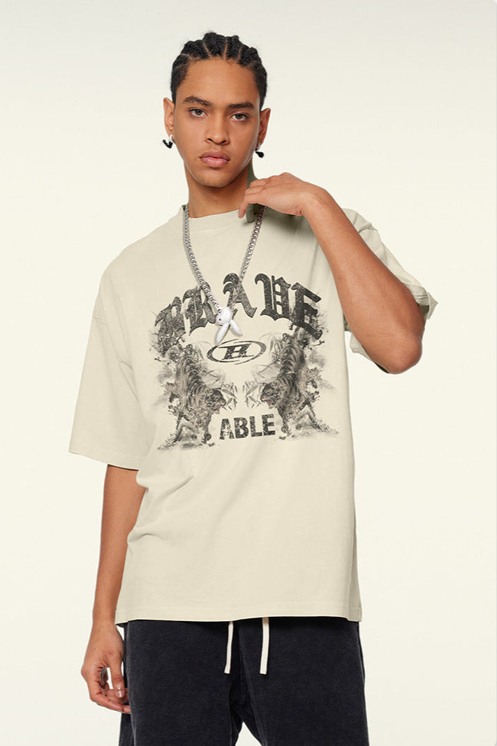 Close-up of model with braided hair touching chain necklace, wearing cream "BRAVE ABLE" graphic t-shirt with tiger motifs against white background