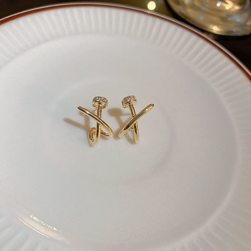 A pair of gold colored crossover hoop earrings with small embedded zirconia, displayed on a white ceramic plate.