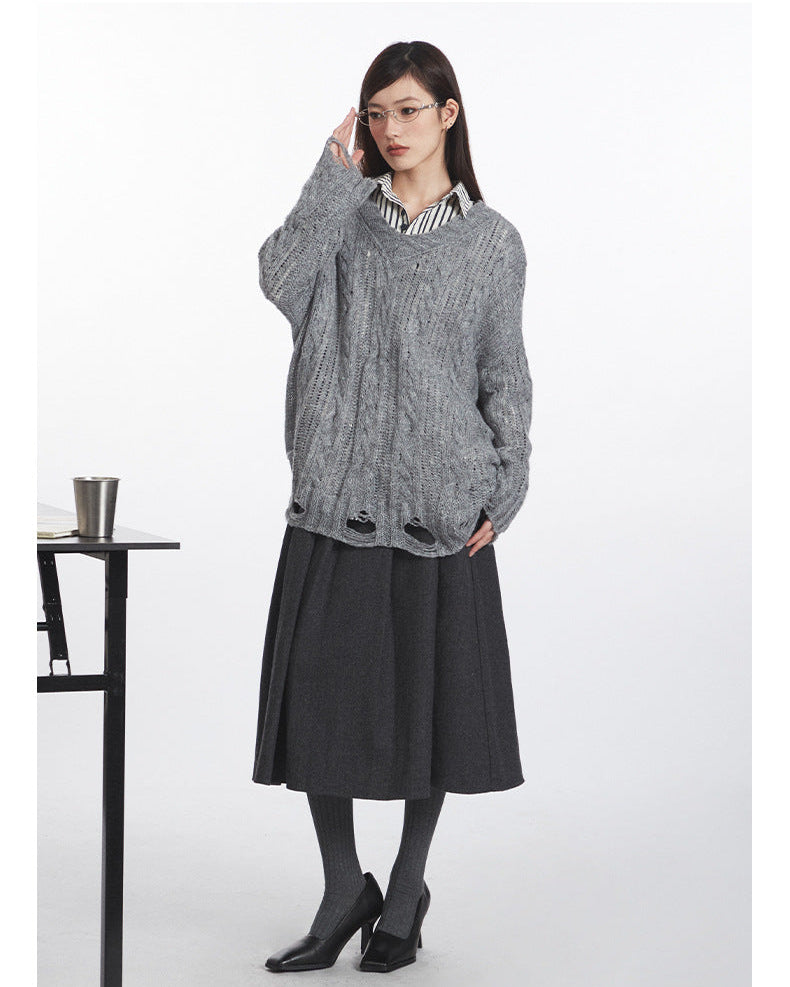 Angled view of grey V-neck sweater highlighting cable-knit pattern and intentionally distressed edges, styled with midi skirt and striped shirt collar