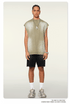 A man wearing a sage green sleeveless muscle tee and black shorts stands against a white background with silver chain necklace and white shoes