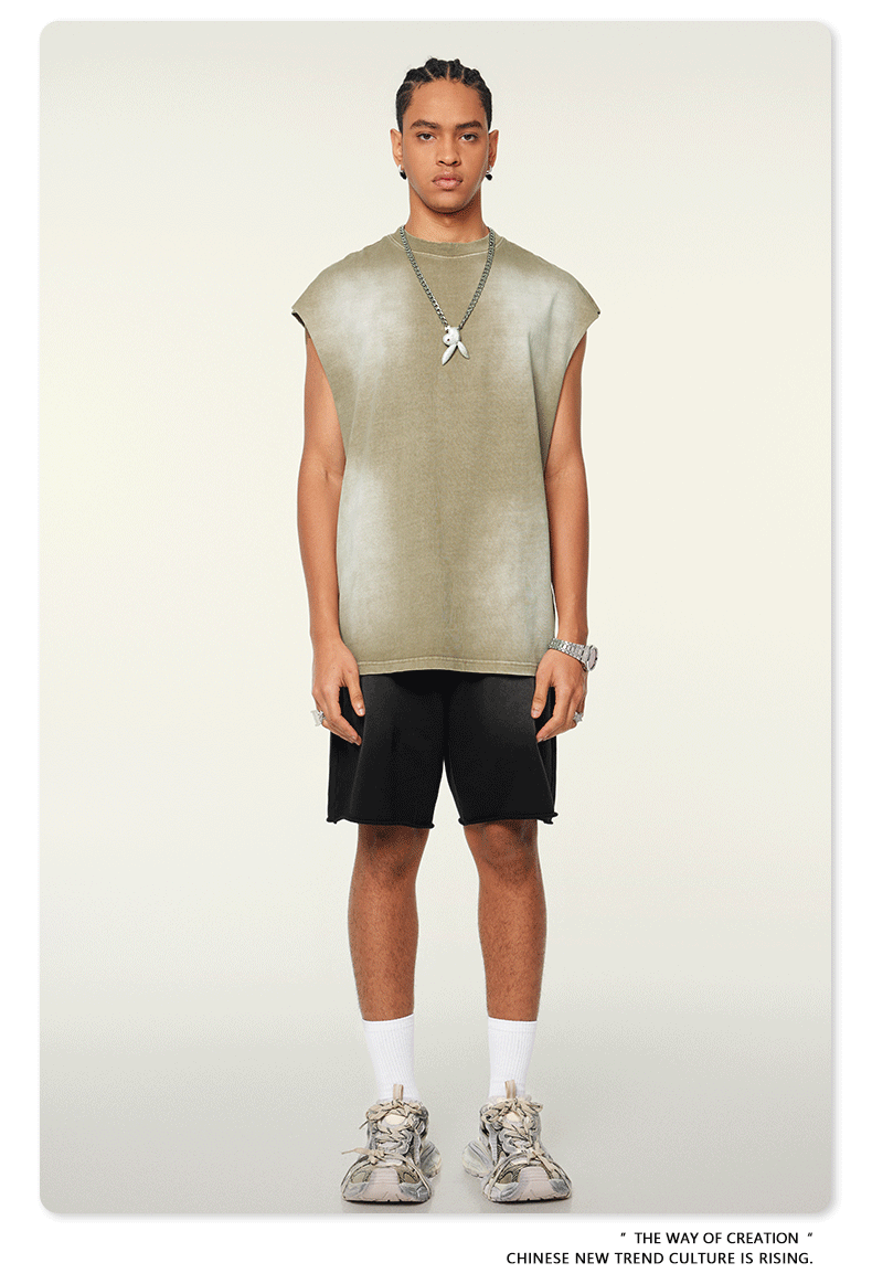 A man wearing a sage green sleeveless muscle tee and black shorts stands against a white background with silver chain necklace and white shoes
