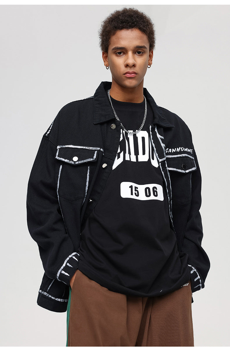 A man is standing against a gray background wearing a black SANHOMME outline denim jacket, graphic tee, and loose brown pants