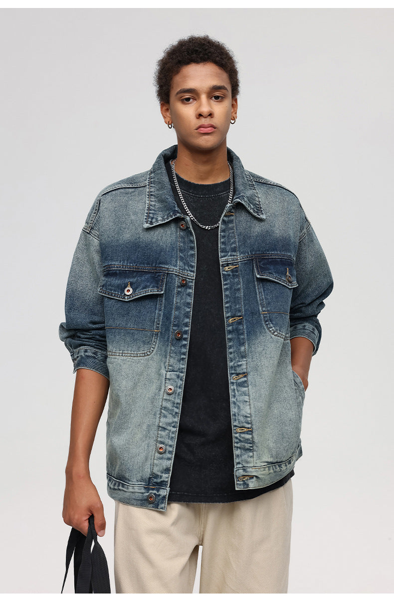 A man is standing front-facing against a gray backdrop. Wearing gradient-wash denim jacket, black tee, silver chain necklace and holding black fabric