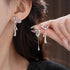 Close-up of crystal-encrusted silver bow earrings on ear, featuring a teardrop pendant on delicate chain and manicured fingers