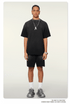 Model with braided hair wearing oversized black t-shirt with silver pendant necklace, matching shorts, white socks and athletic shoes against white background