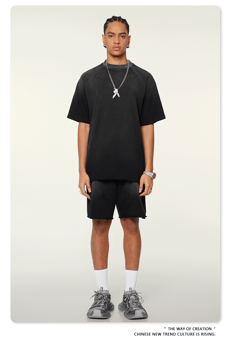 Model with braided hair wearing oversized black t-shirt with silver pendant necklace, matching shorts, white socks and athletic shoes against white background