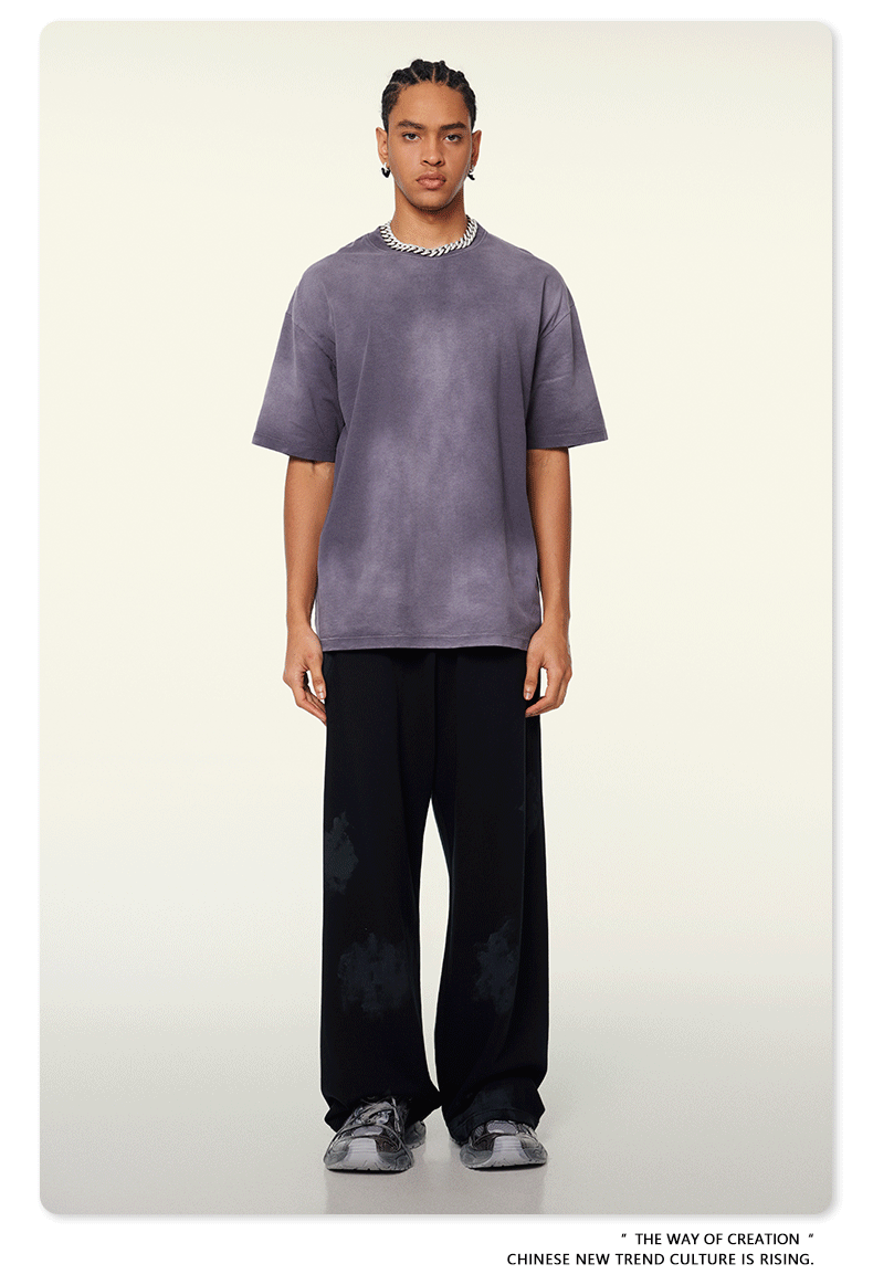 Model with braided hair wearing slate blue t-shirt and black wide-leg pants with silver chain necklace against cream background