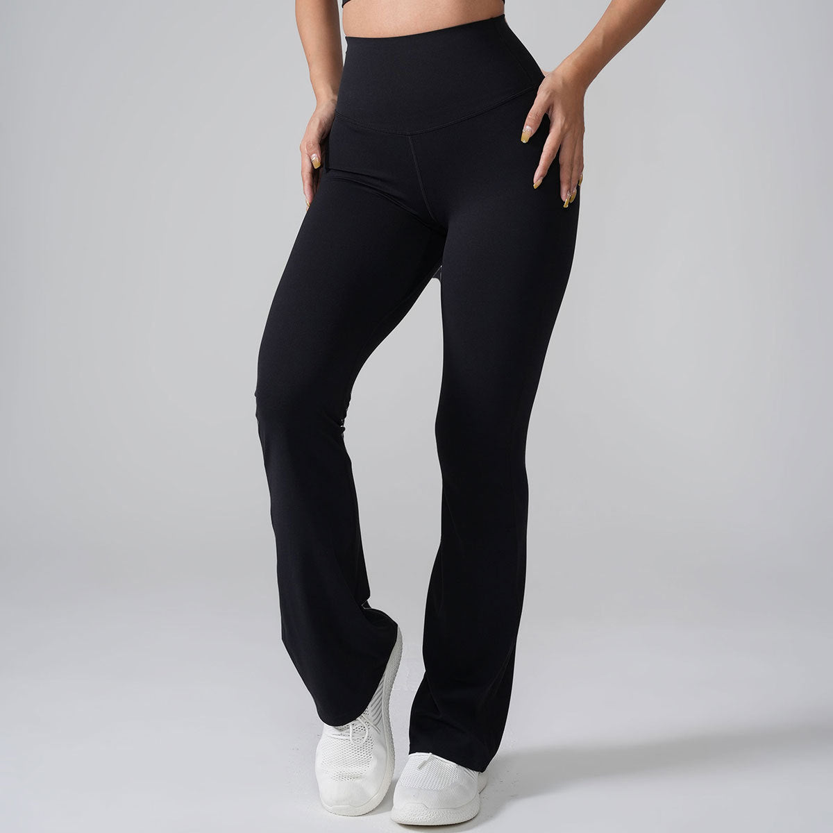 A model wearing black high-waisted flared yoga pants with white sneakers, shown full-length against gray backdrop