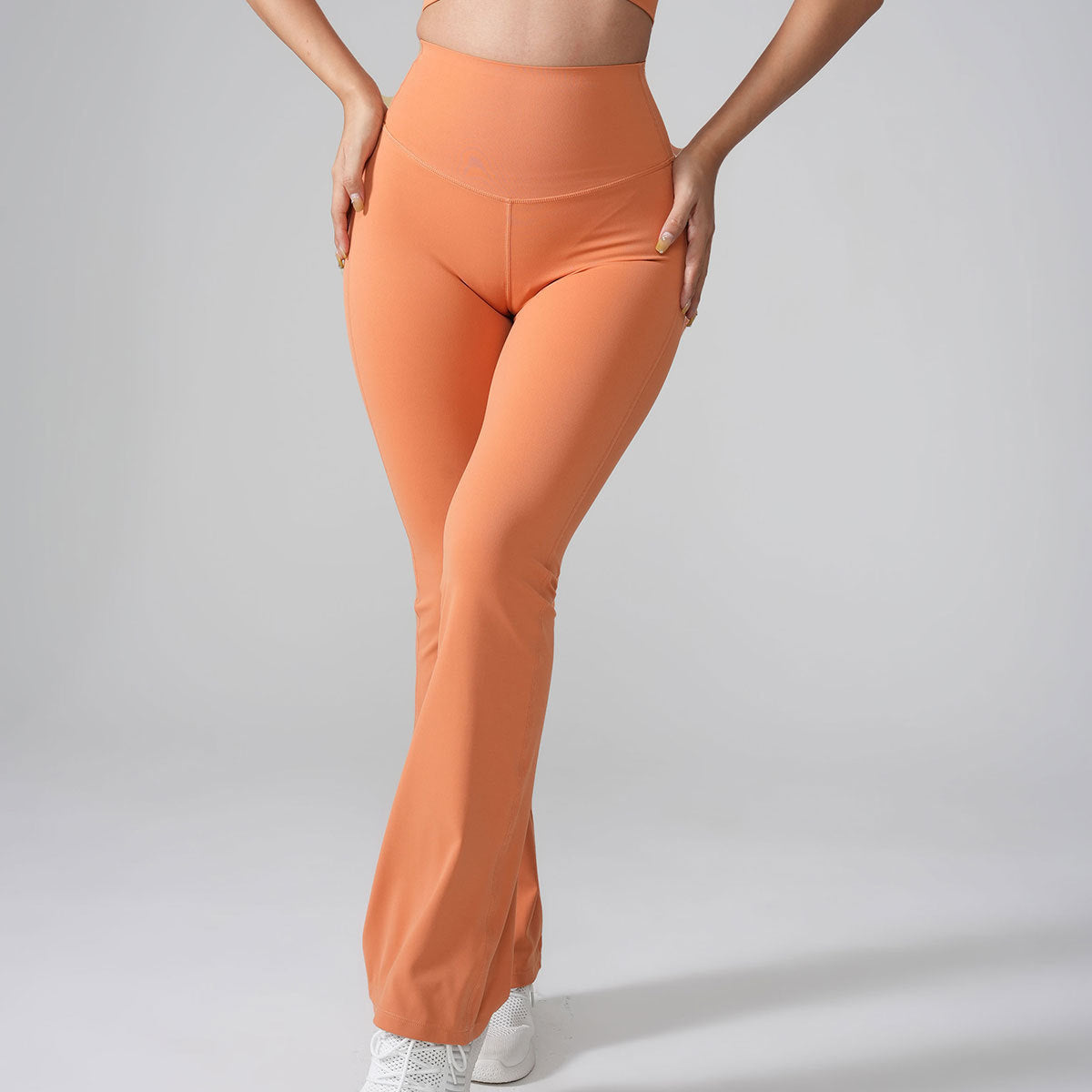 A model wearing coral-colored high-waisted flared yoga pants with white athletic shoes, posed against a neutral backdrop