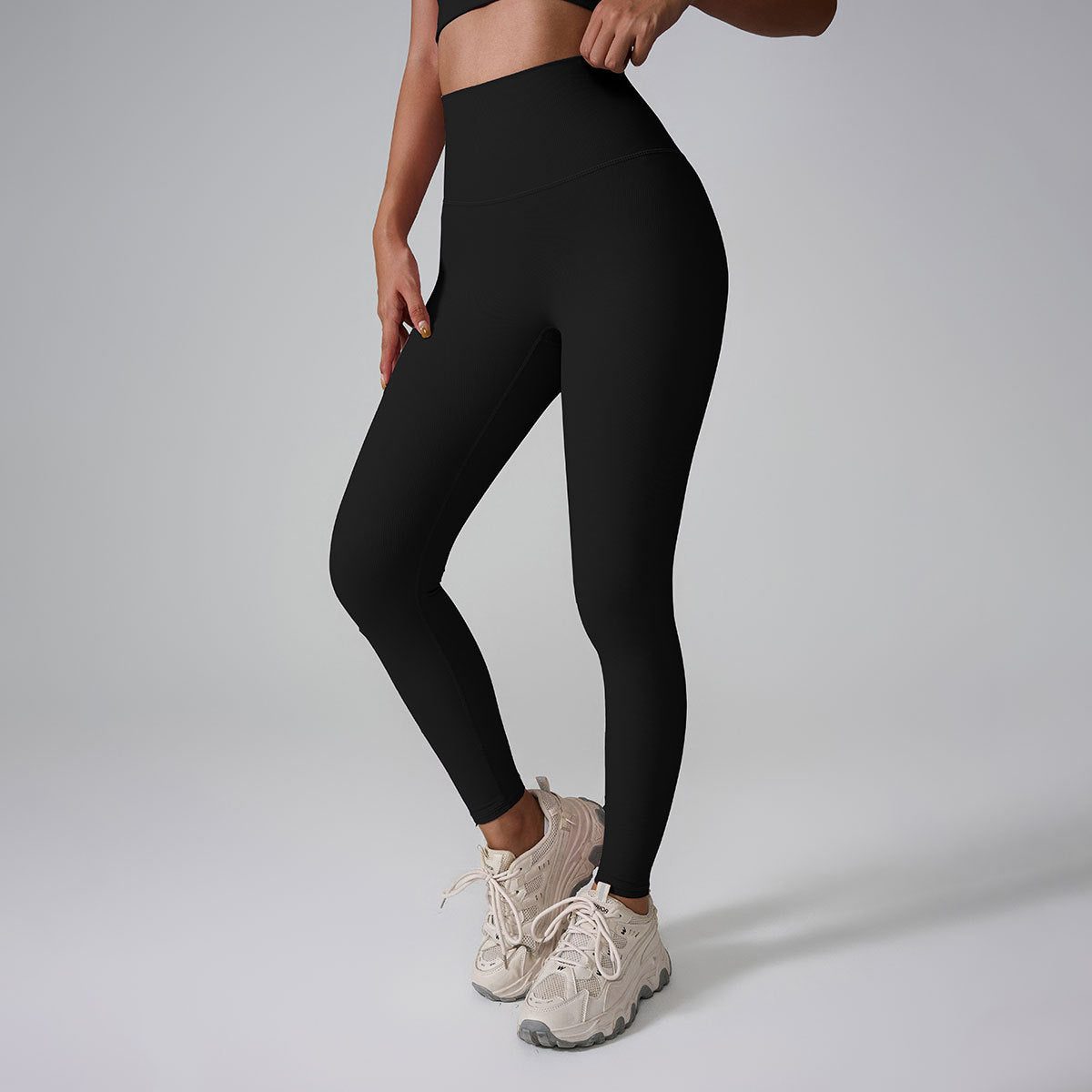A model wearing black high-waisted workout leggings and crop top, standing against gray backdrop with cream athletic shoes