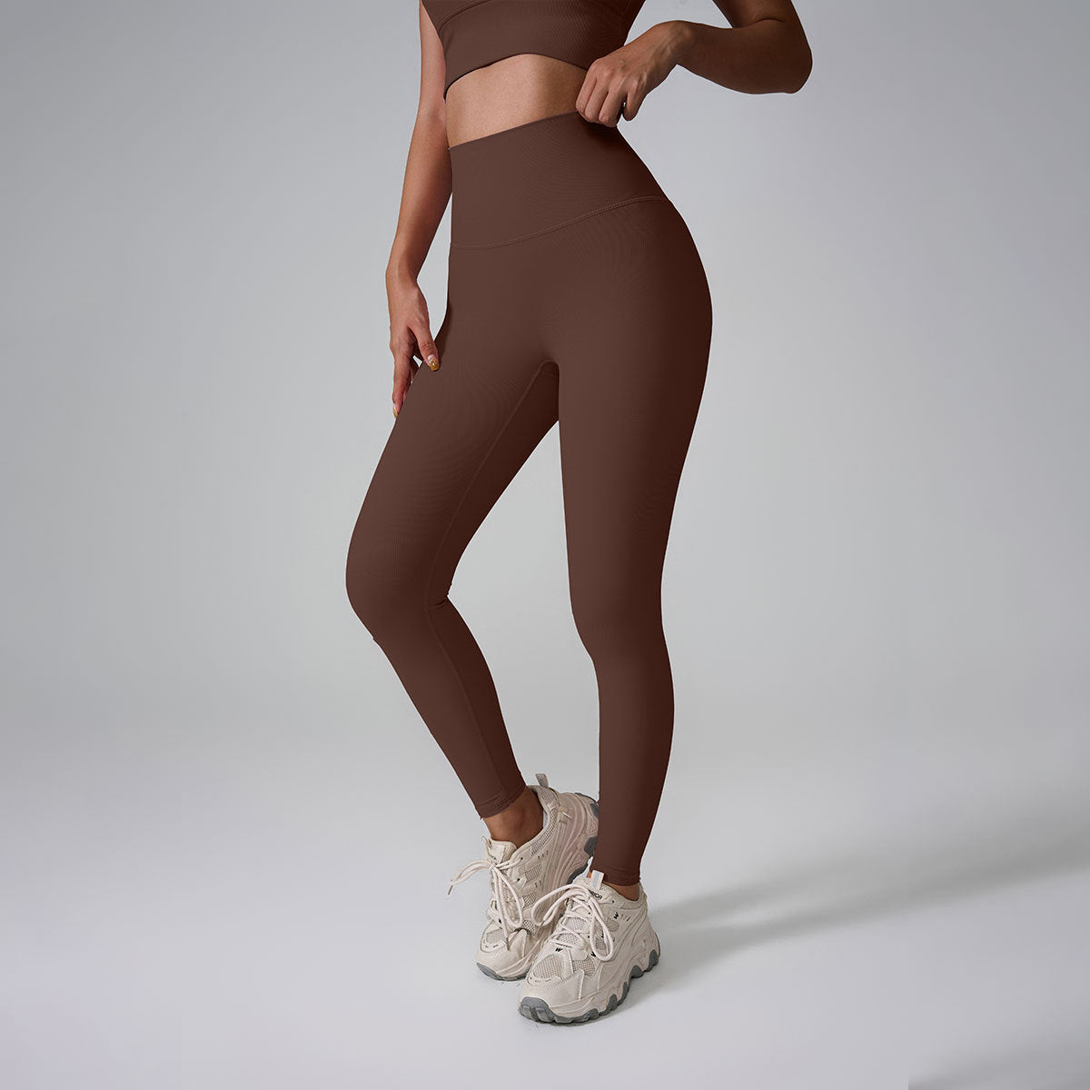 A model wearing chocolate brown high-waisted workout leggings and matching sports bra against gray backdrop