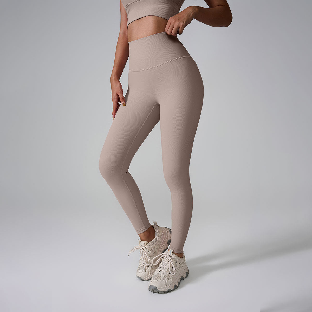 A model wearing beige high-waisted workout leggings and matching crop top against gray studio background