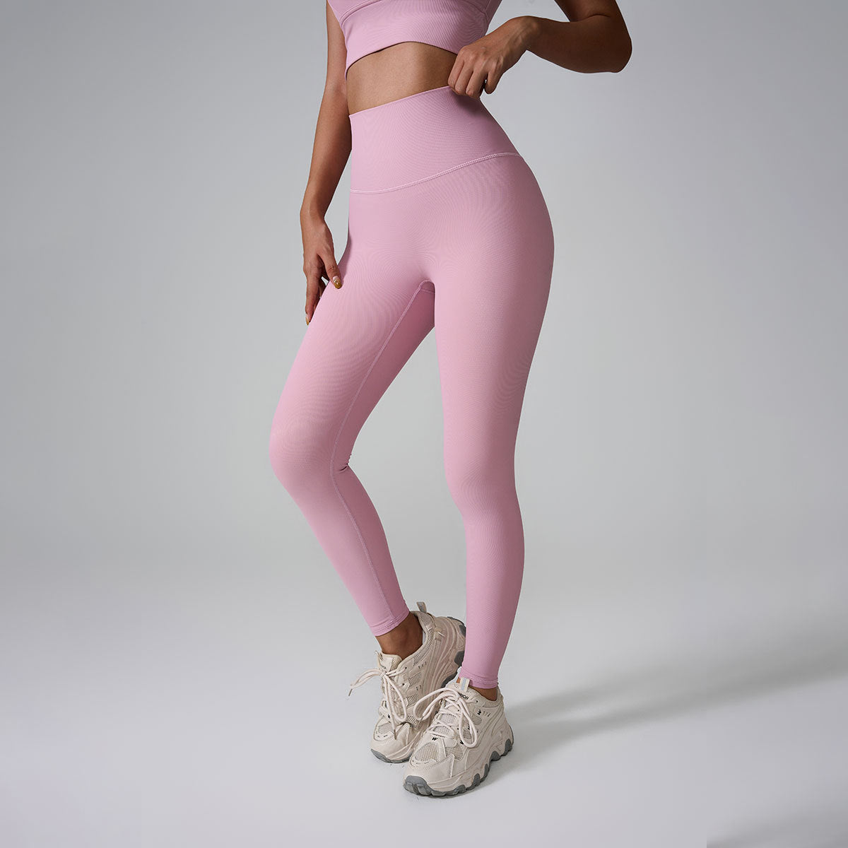 A model wearing pink high-waisted workout leggings and matching crop top, standing with hands on hips against gray background