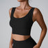 A woman wearing a black ribbed scoop neck sports bra with wide straps and supportive seam details against a minimalist gray background