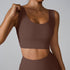 A woman wearing a chocolate brown ribbed scoop neck sports bra while adjusting strap with one hand, paired with matching leggings against gray backdrop