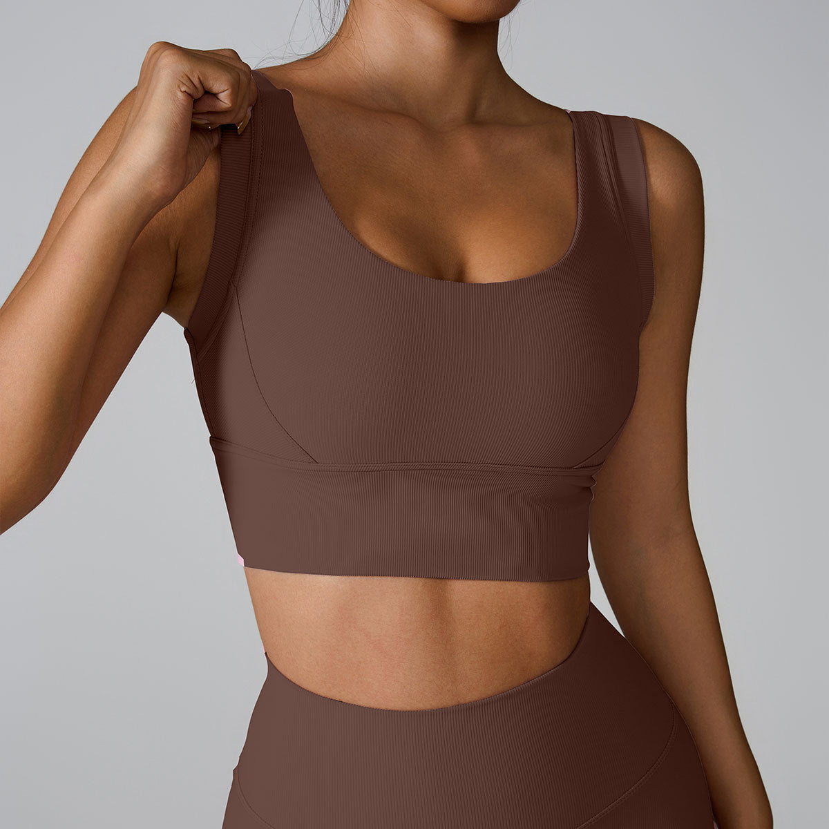 A woman wearing a chocolate brown ribbed scoop neck sports bra while adjusting strap with one hand, paired with matching leggings against gray backdrop