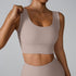 A woman wearing a beige ribbed scoop neck sports bra while adjusting strap with one hand, showing seamless design against a neutral gray backdrop