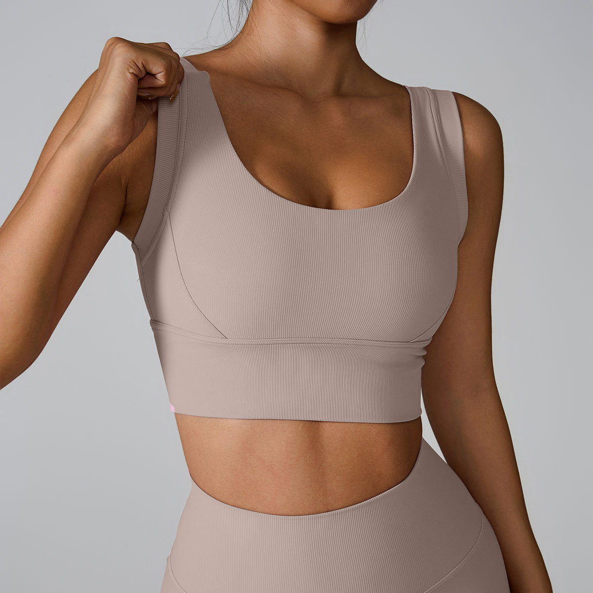 A woman wearing a beige ribbed scoop neck sports bra while adjusting strap with one hand, showing seamless design against a neutral gray backdrop