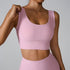 A woman wearing a light pink ribbed scoop neck sports bra with wide straps while adjusting strap with one hand, paired with matching high-waisted bottoms