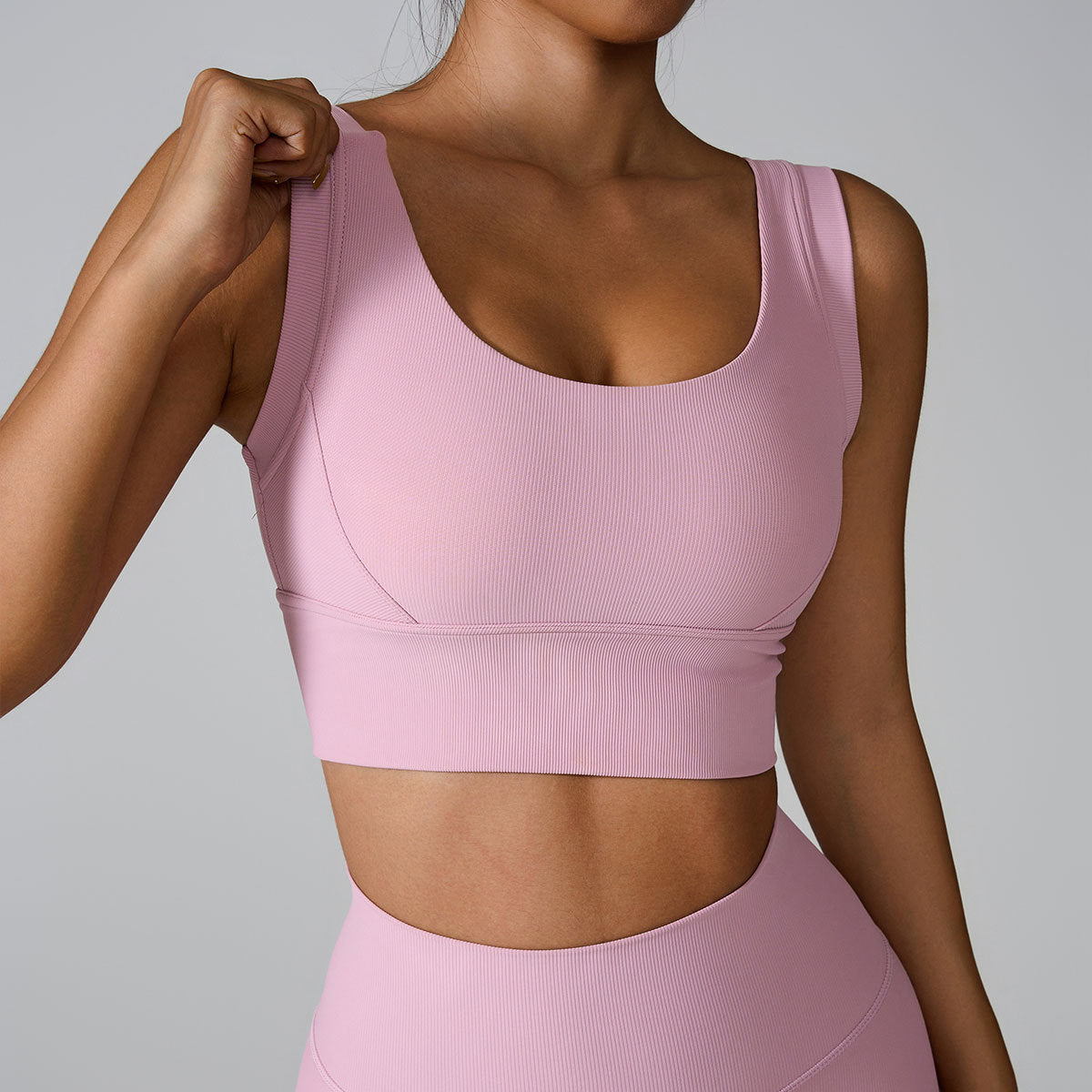 A woman wearing a light pink ribbed scoop neck sports bra with wide straps while adjusting strap with one hand, paired with matching high-waisted bottoms