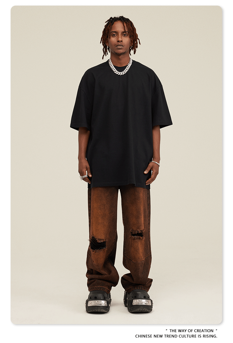 Model with dreadlocks wearing black oversized t-shirt with brown distressed jeans and black footwear against beige background