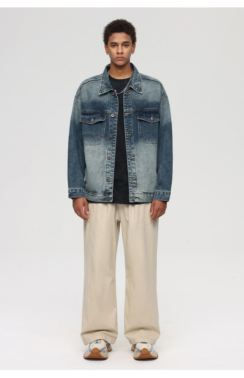 A man is posing straight-on against a gray background. Wearing oversized faded denim trucker jacket, black t-shirt, beige wide-leg pants and athletic sneakers