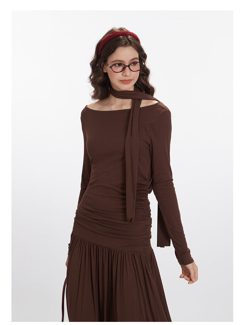 Brown jersey maxi dress featuring off-shoulder neckline with integrated neck scarf, ruched side panels, long fitted sleeves, and full-length gathered skirt