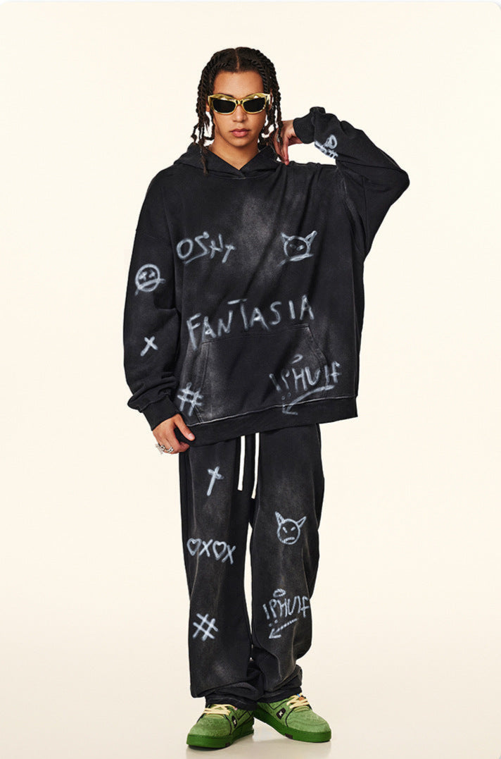 Model in black graffiti hoodie and sweatpants with white painted symbols, adjusting gold sunglasses against neutral background