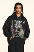A man is standing with hands in pockets wearing a black graphic hoodie featuring artistic text and skull imagery with silver chain necklace