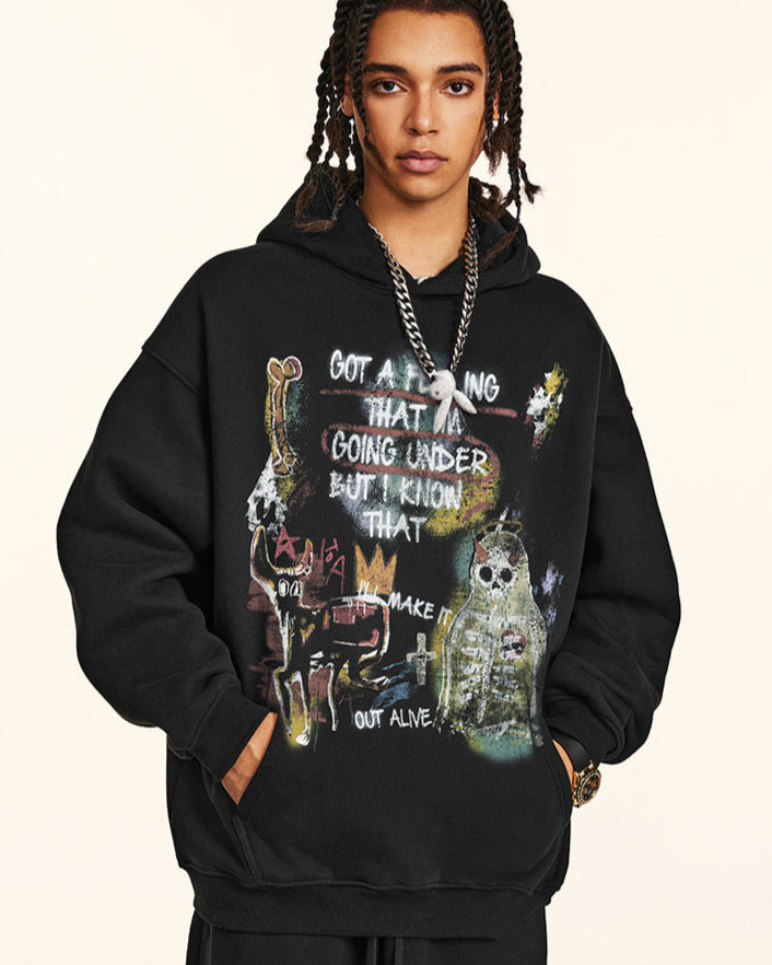 A man is standing with hands in pockets wearing a black graphic hoodie featuring artistic text and skull imagery with silver chain necklace