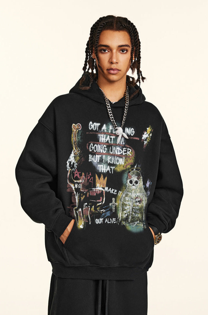 A man is standing with hands in pockets wearing a black graphic hoodie featuring artistic text and skull imagery with silver chain necklace