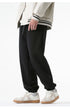 A model is wearing black jogger sweatpants with a relaxed fit, elastic cuffs, and a small embroidered logo, paired with a cream varsity jacket and white sneakers