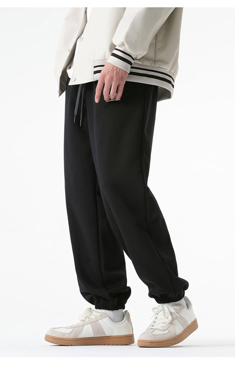 A model is wearing black jogger sweatpants with a relaxed fit, elastic cuffs, and a small embroidered logo, paired with a cream varsity jacket and white sneakers