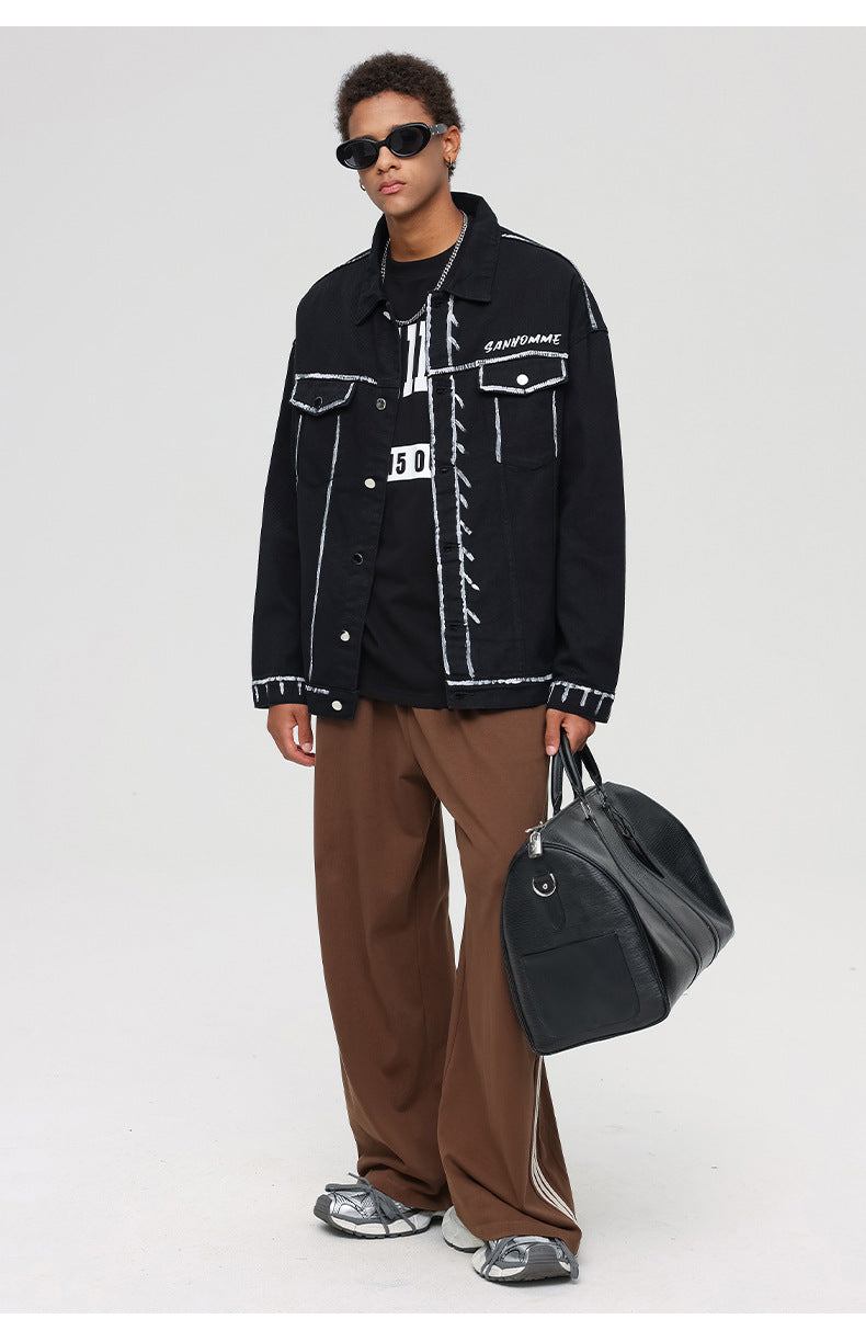 A man is posing with sunglasses and a black duffel bag. Wearing black outline denim jacket, brown pants and silver sneakers on gray backdrop