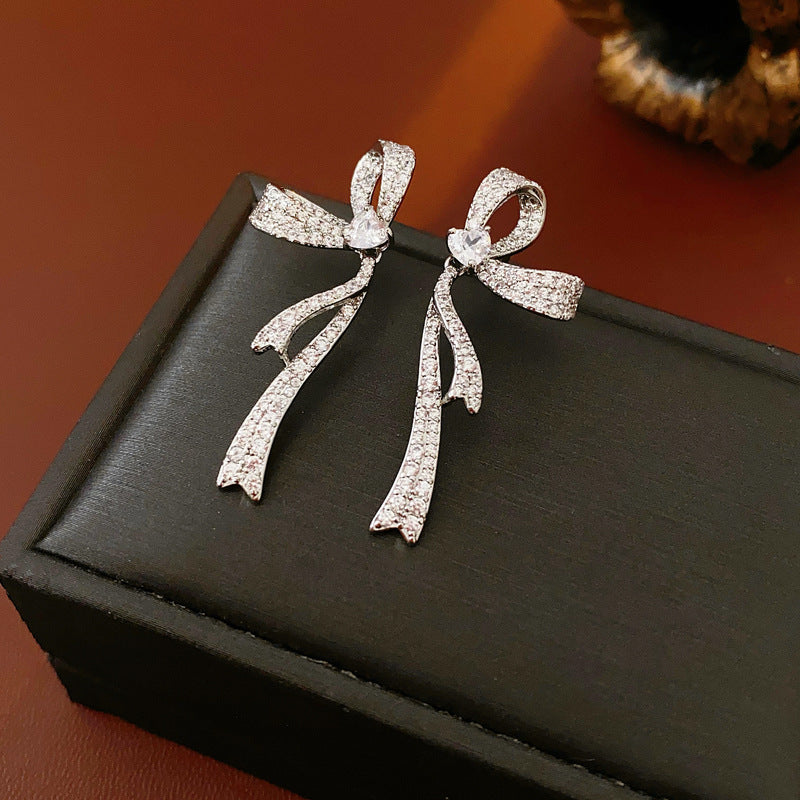 Pair of silver bow earrings with pavé crystal details displayed on black jewelry box against brown background