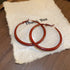 A pair of large gold hoop earrings with a wavy red enamel design, resting on a beige fabric jewelry case