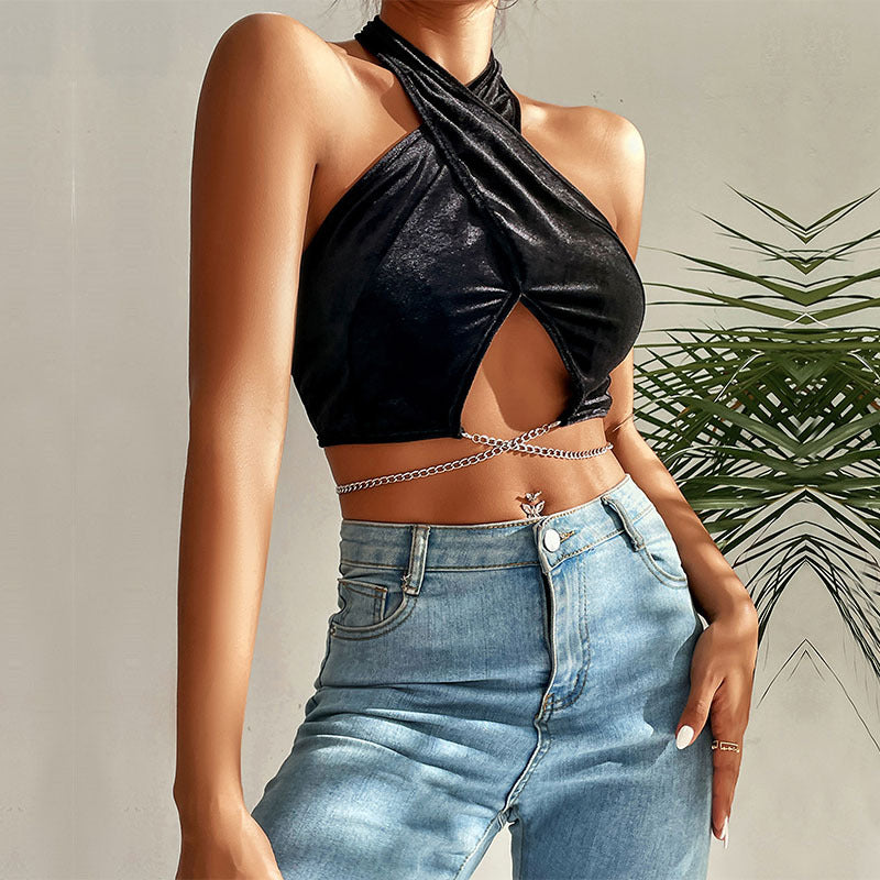 Close-up of black velvet halter top featuring criss-cross neckline, decorative belly chain, and high-waisted denim jeans against greenery