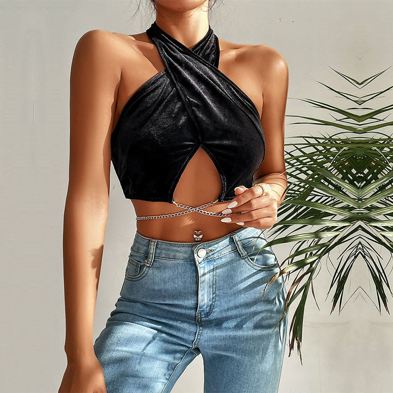 A model wearing a black velvet halter crop top with crossed front design, silver waist chain, and light blue jeans, styled with palm leaves backdrop