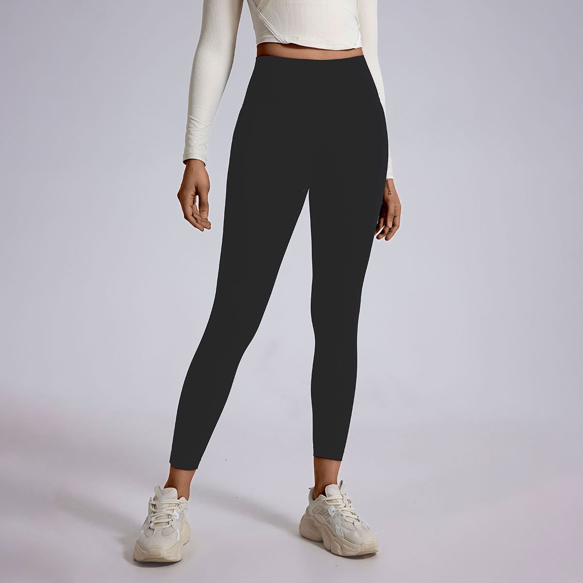 A model in black high-waisted compression leggings and white cropped top, wearing white athletic sneakers, studio lighting