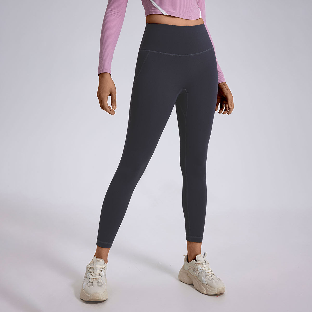 A model wearing black ankle-length leggings with pink long-sleeve crop top and white chunky sneakers, gray studio background