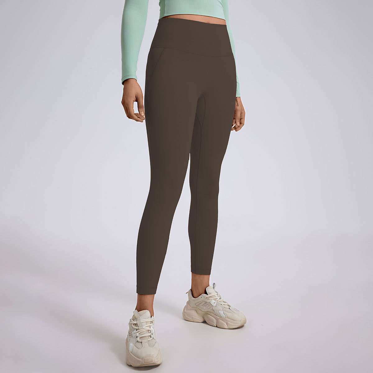 A model wearing dark brown athletic leggings with mint green crop top and cream colored chunky sneakers, gray backdrop