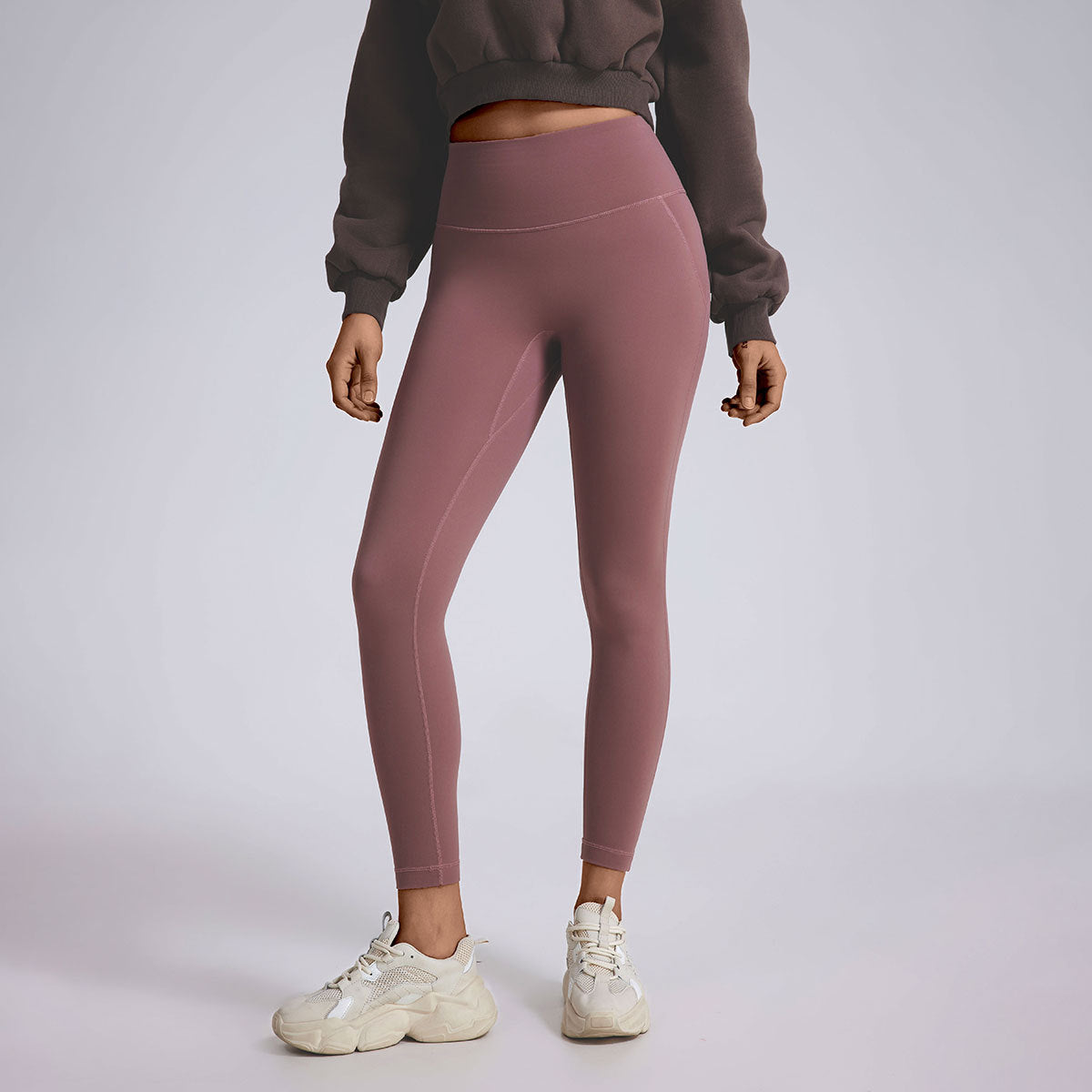 A model wearing mauve high-waisted leggings, brown cropped sweatshirt, and cream chunky sneakers, photographed against gray backdrop