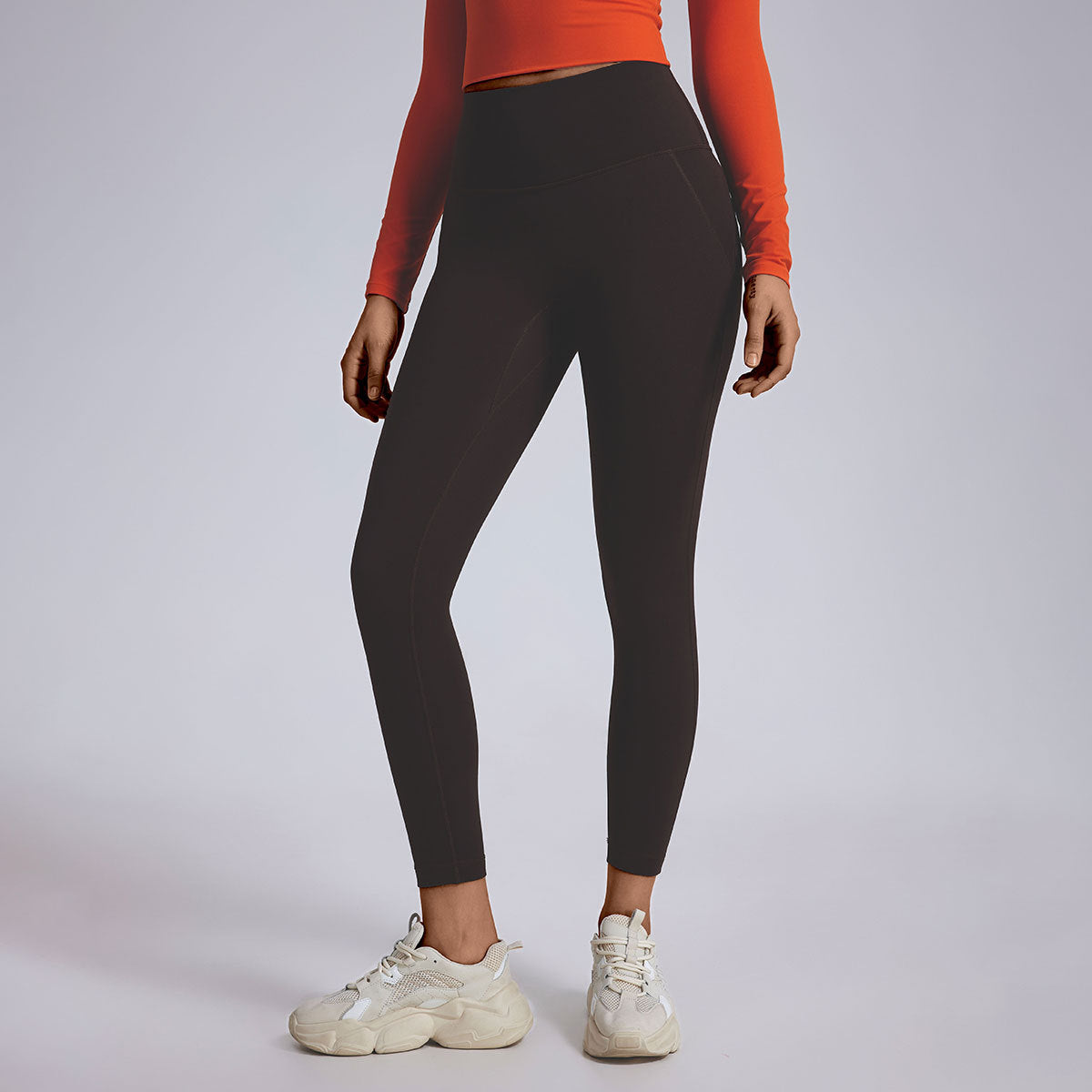 A model in black ankle-length leggings with orange long-sleeve crop top and white athletic sneakers, studio lighting