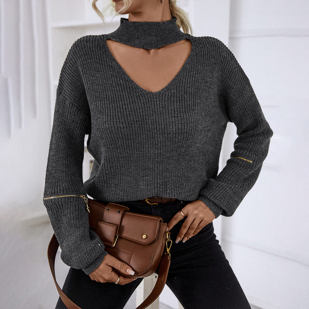Woman wearing gray knit sweater with neck cutout, gold sleeve zippers, black jeans, and brown leather bag against white wall