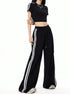 A woman is standing with hand on hip modeling a sporty black crop top with white stripe sleeves and coordinating black wide-leg pants