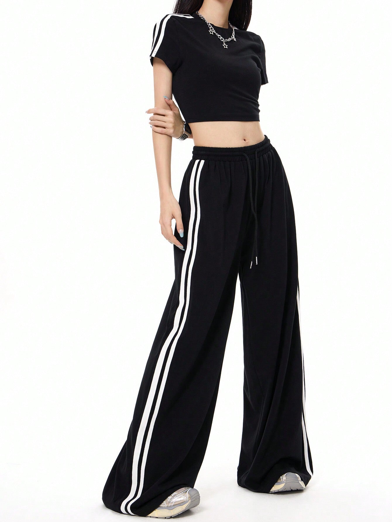A woman is standing with hand on hip modeling a sporty black crop top with white stripe sleeves and coordinating black wide-leg pants