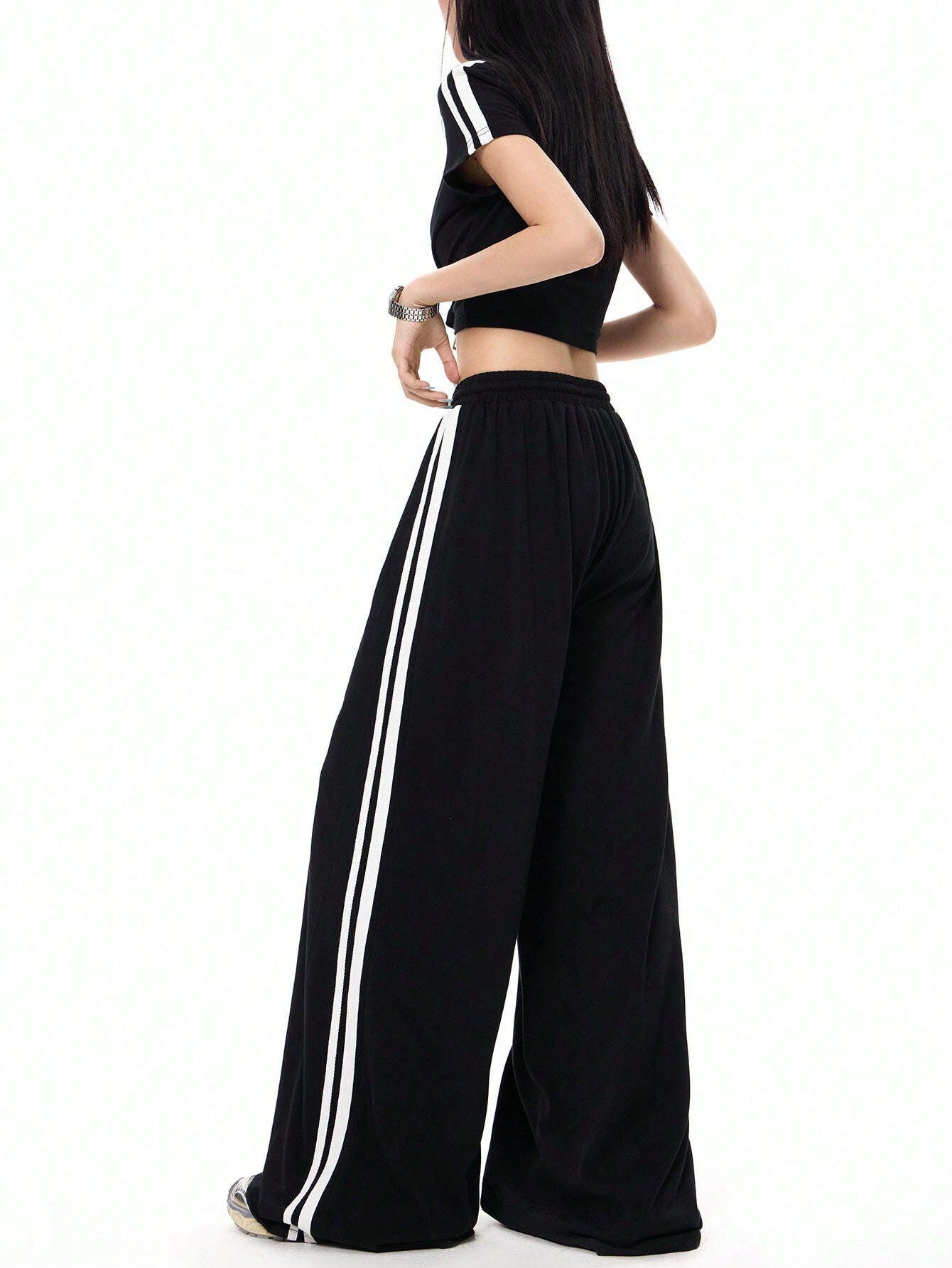 A woman is posing in a side view wearing black wide-leg track pants with white side stripes and a matching black crop top with athletic styling