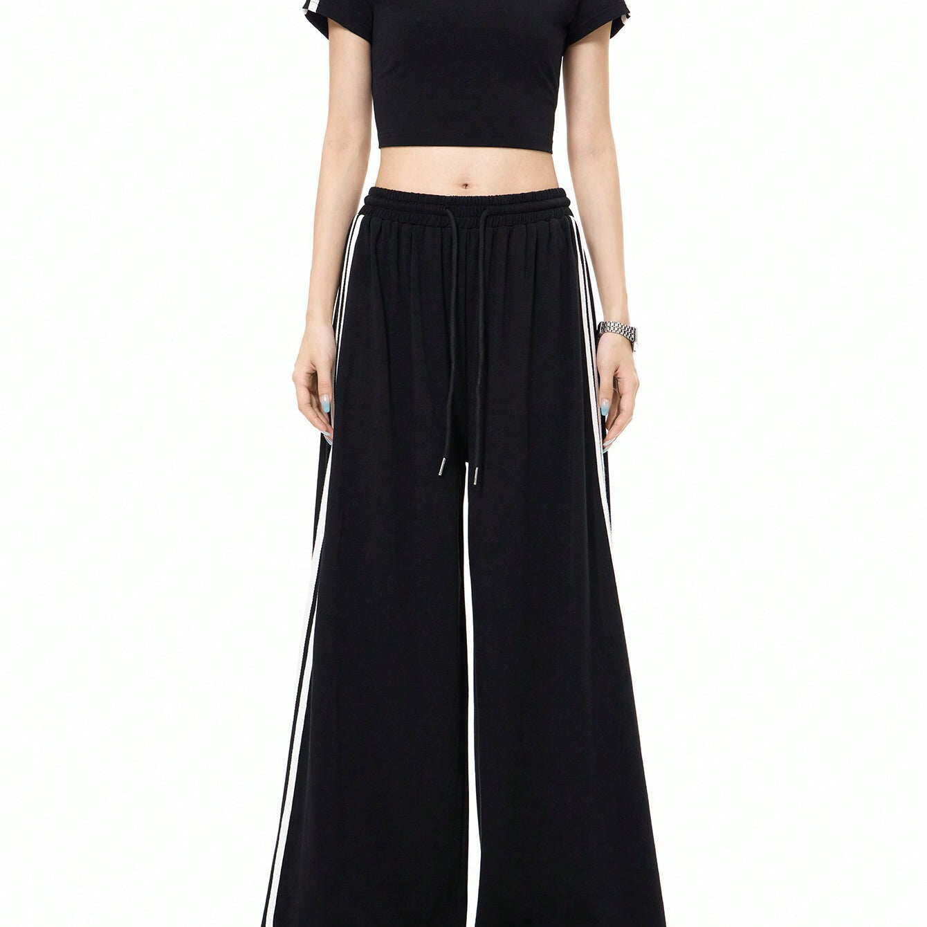 A woman is standing in a black athleisure two-piece set with white side stripes, featuring a crop top and wide-leg pants with drawstring waist