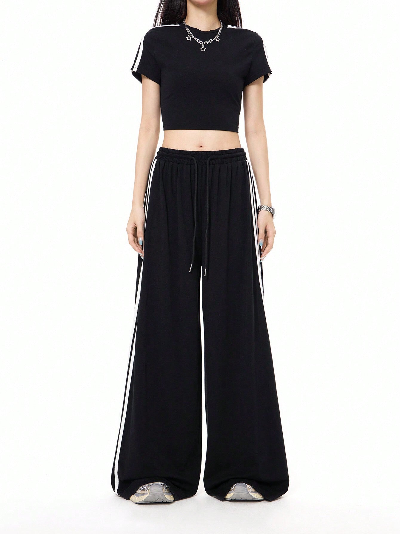 A woman is standing in a black athleisure two-piece set with white side stripes, featuring a crop top and wide-leg pants with drawstring waist