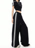 A woman is standing in profile view showcasing a black athletic two-piece outfit with white side stripes and ultra-wide palazzo-style pants